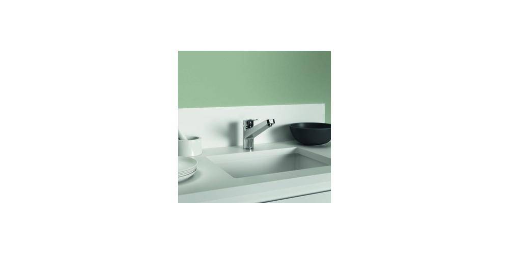 Minimalistische Küchenspüle mit Chromarmatur, weißer Arbeitsplatte und einer schwarzen Schüssel im Hintergrund vor einer blassgrünen Wand.