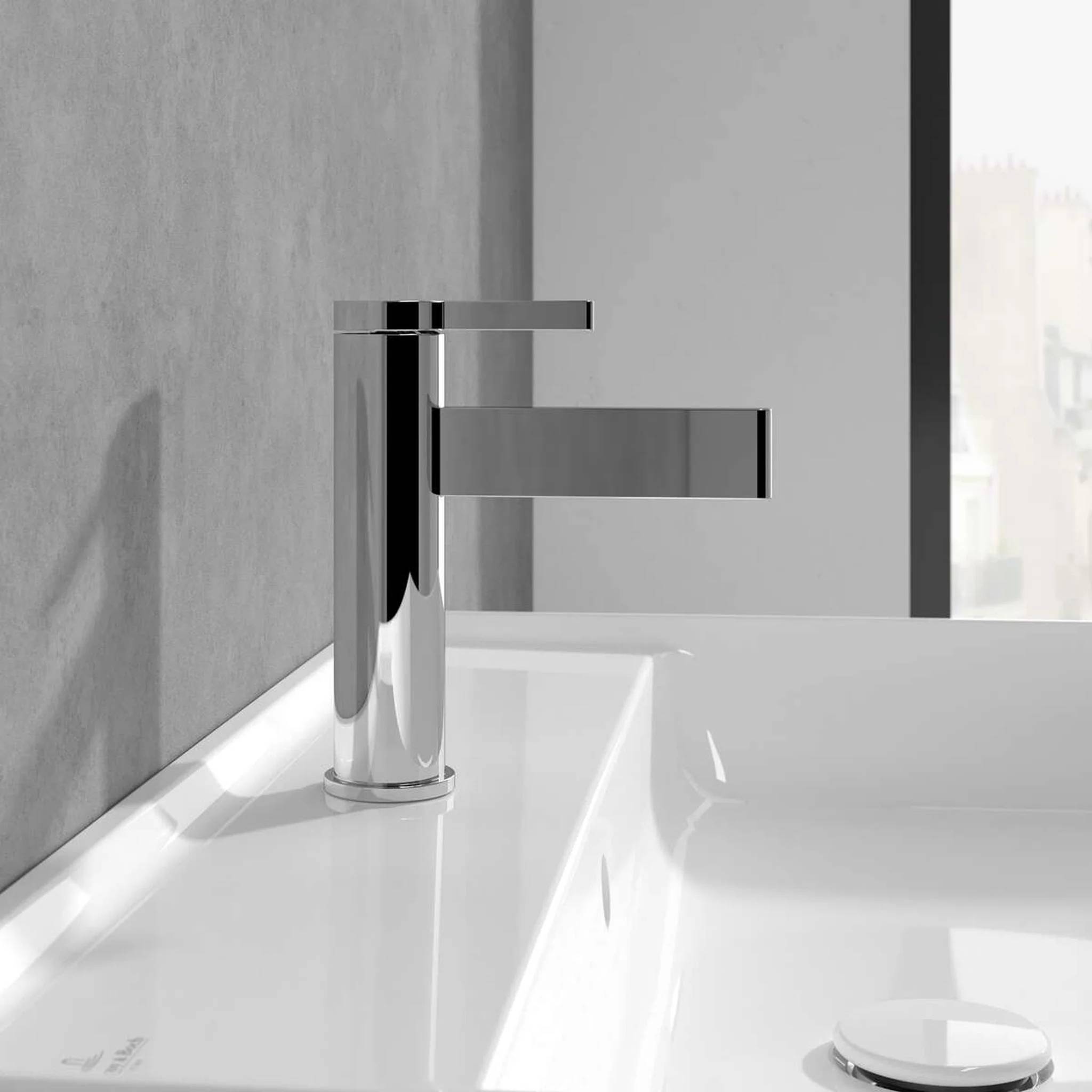 Moderner Wasserhahn aus Chrom im Badezimmer, das auf einem weißen Waschbecken vor einer grauen Wand und einem Spiegel montiert ist.