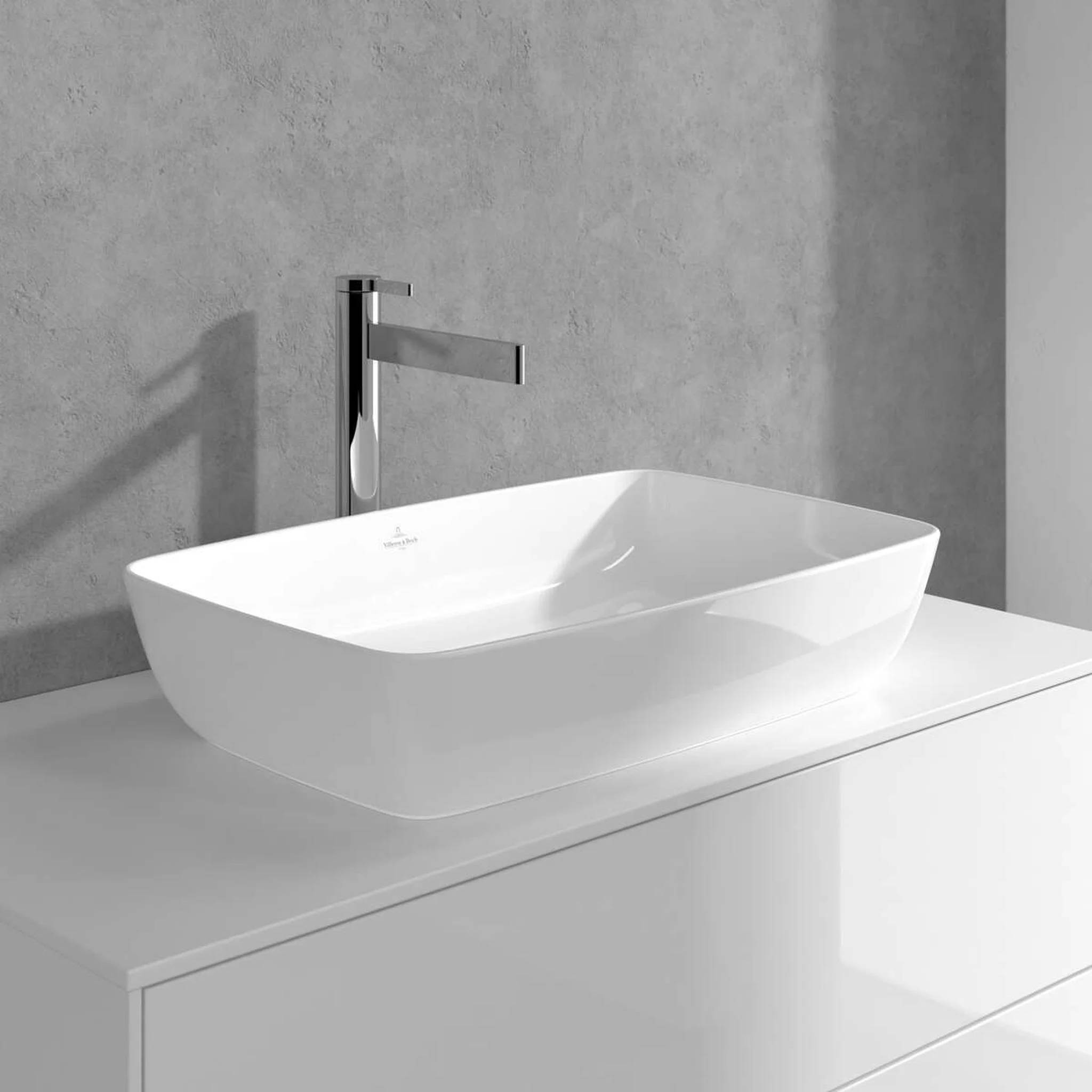 Modernes weißes Waschbecken aus Keramik mit verchromtem Wandarmatur auf einer hellen Arbeitsplatte vor einer grauen Wand.