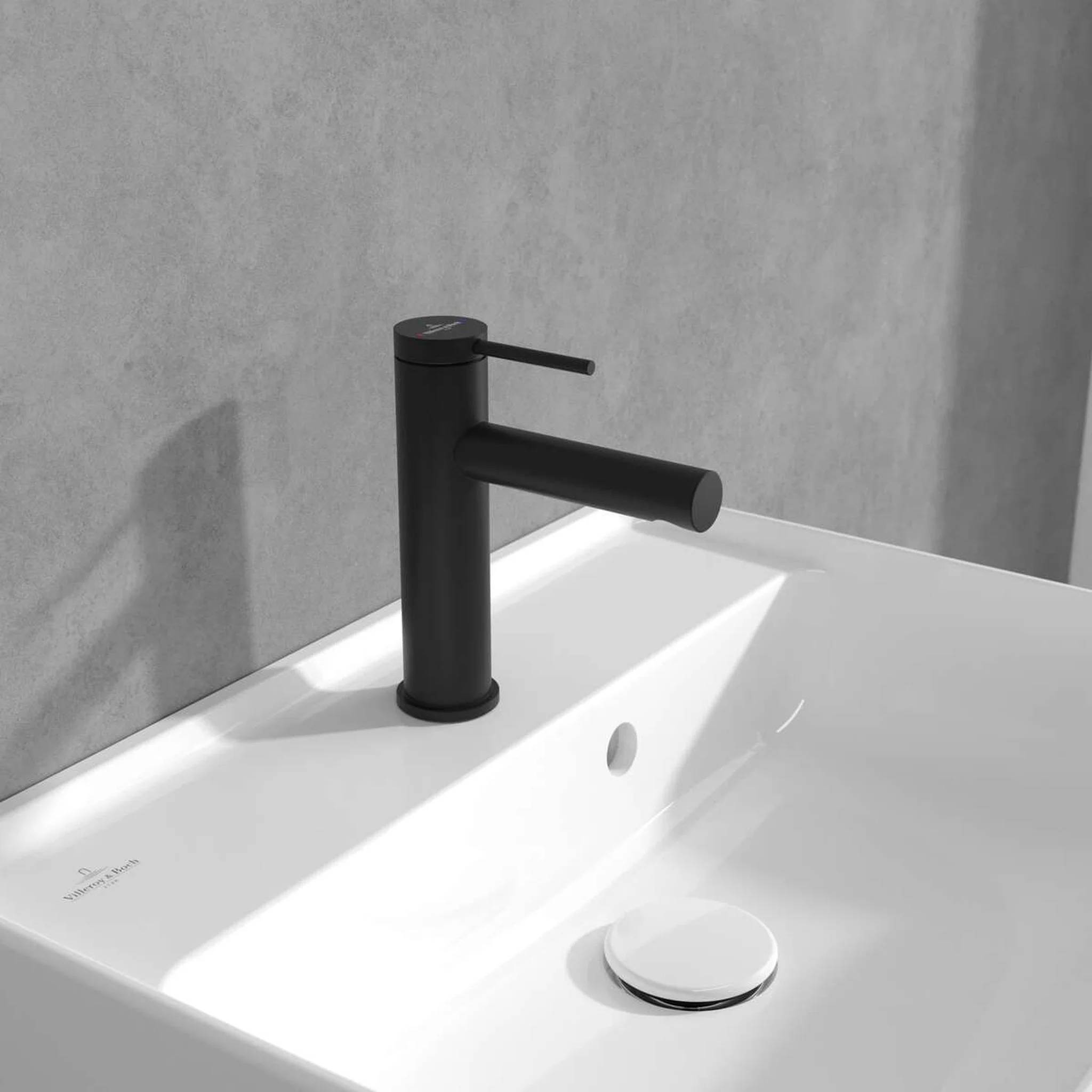 Moderner schwarzer Badezimmerhahn über einem weißen Waschbecken mit passendem Abflussstöpsel und minimalistischem grauem Wandhintergrund.