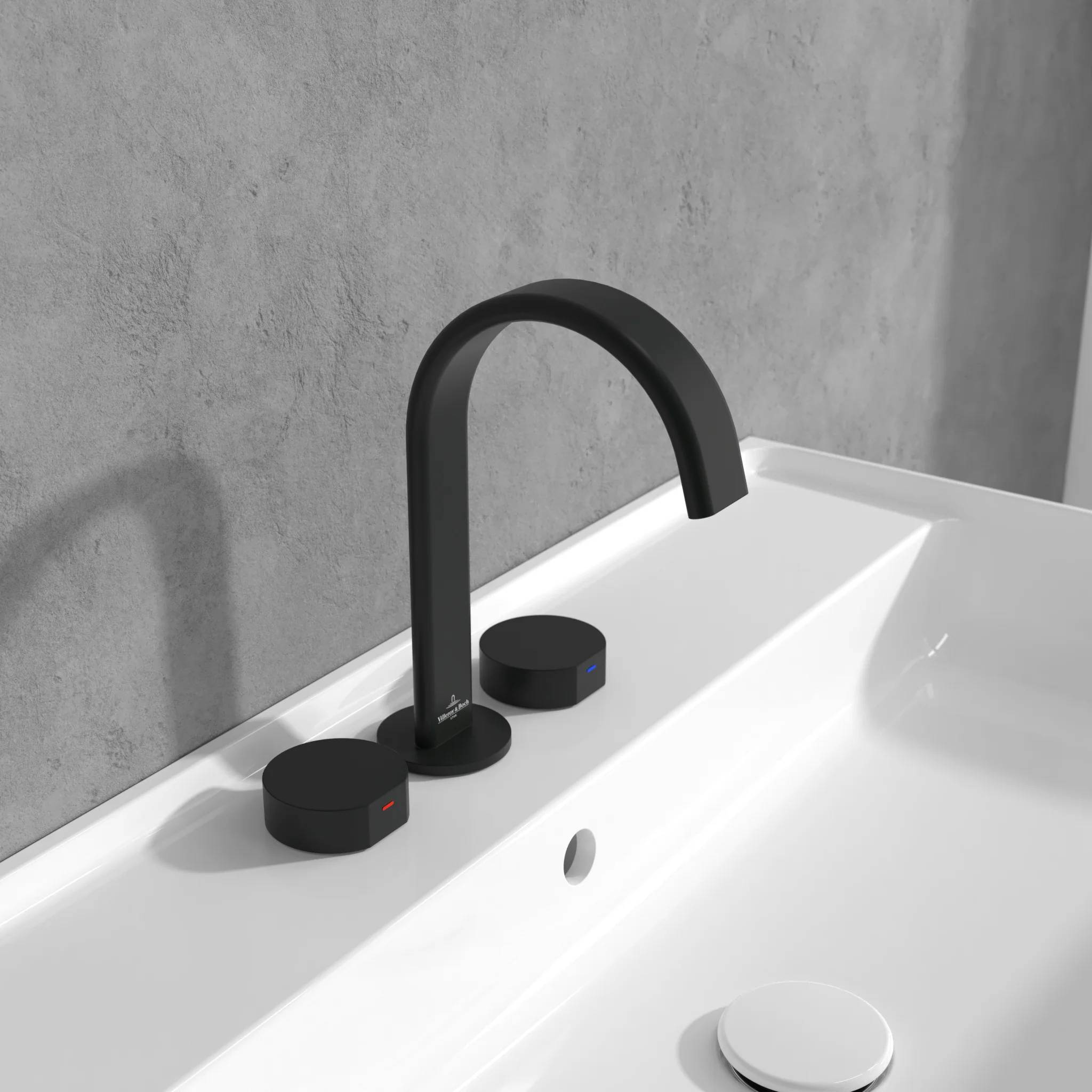 Schwarzer matter Wasserhahn mit zwei runden Bediengriffen auf einem weißen Waschbecken vor einer grauen Wand.