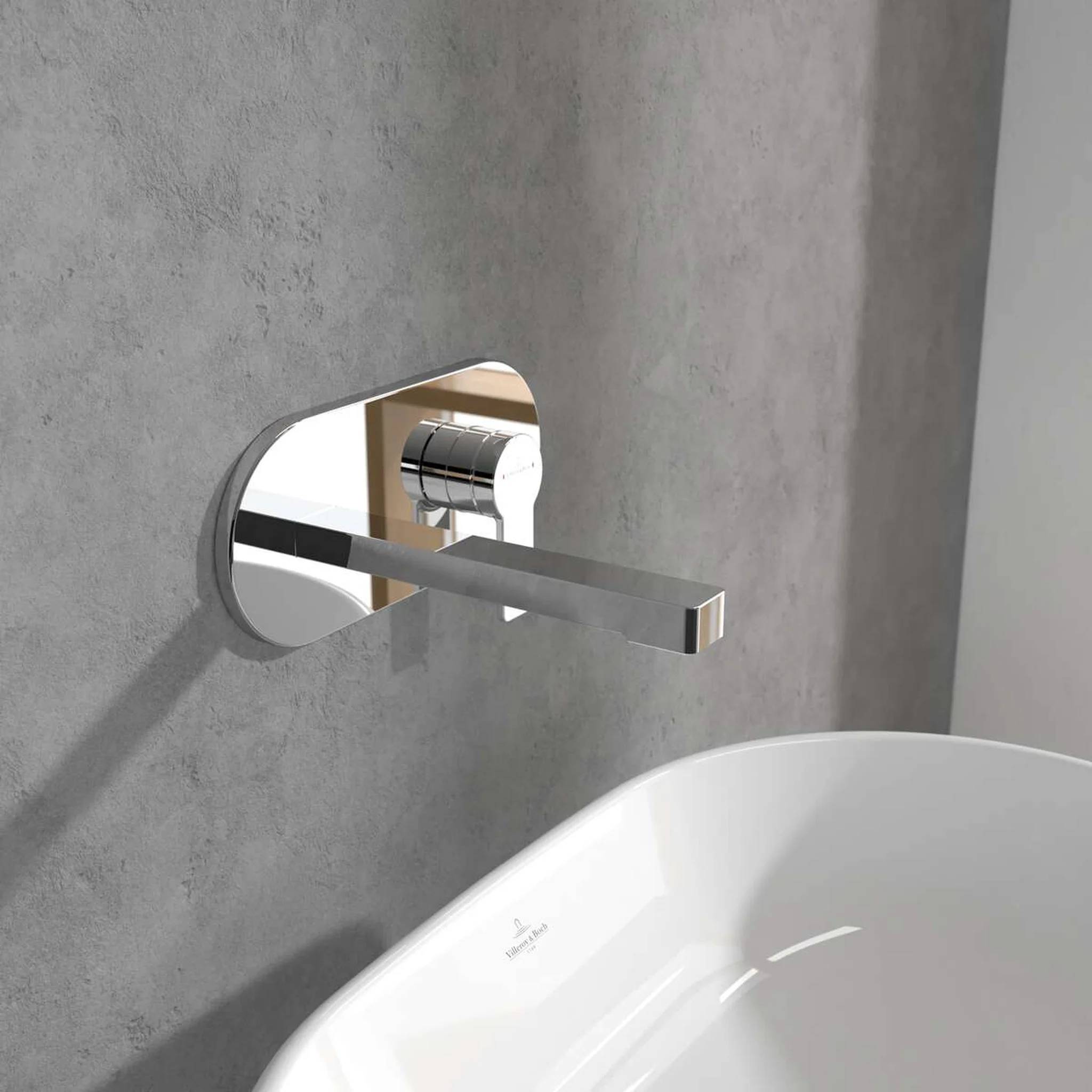 Moderner an der Wand montierter Badezimmerhahn mit langem verchromtem Auslauf über einem weißen Waschbecken, vor einer grau aussehenden Betonwand.
