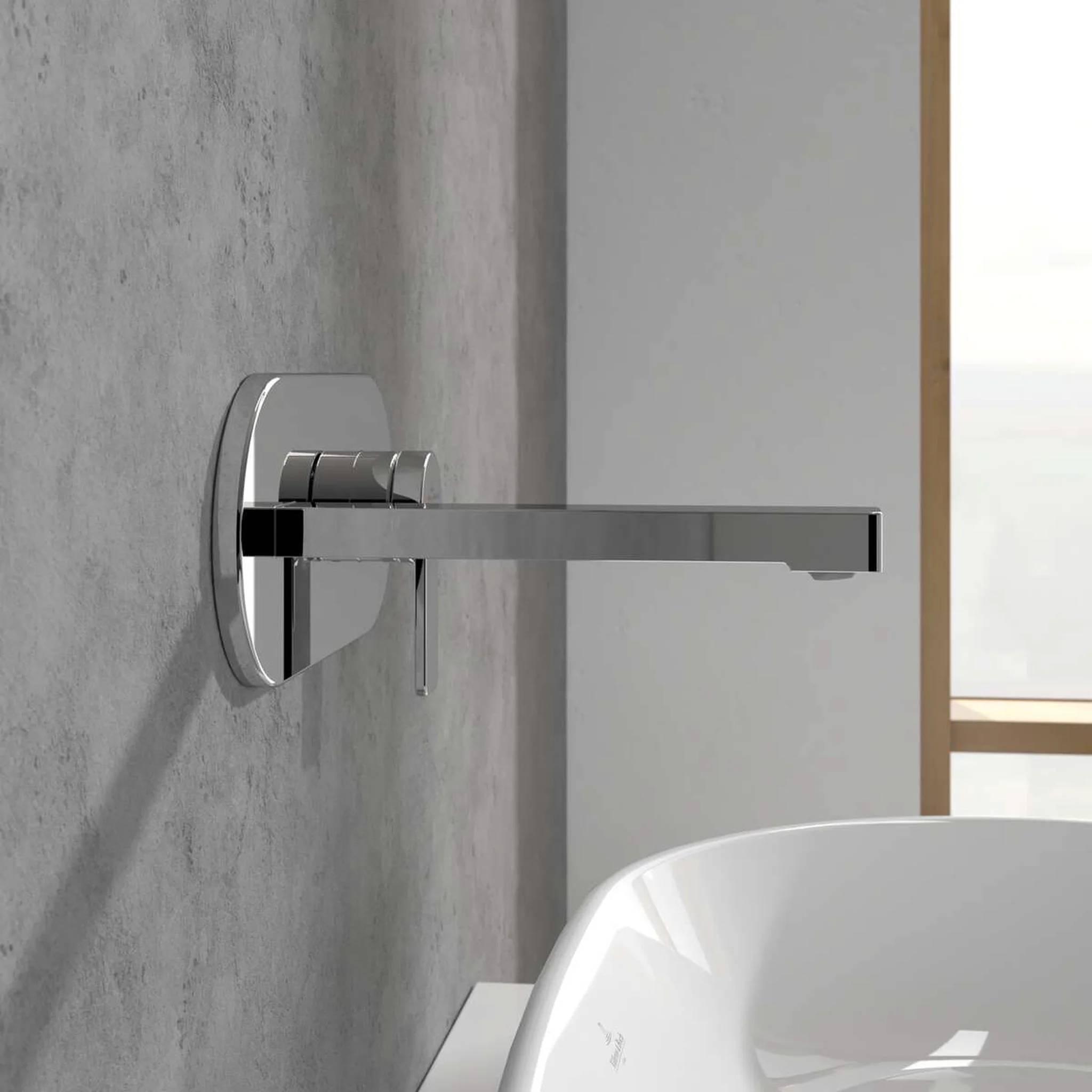 Moderner, wandmontierter Badezimmerwasserhahn aus Chrom, der sich über einem weißen Waschbecken vor einer grau gefliesten Wand erstreckt.