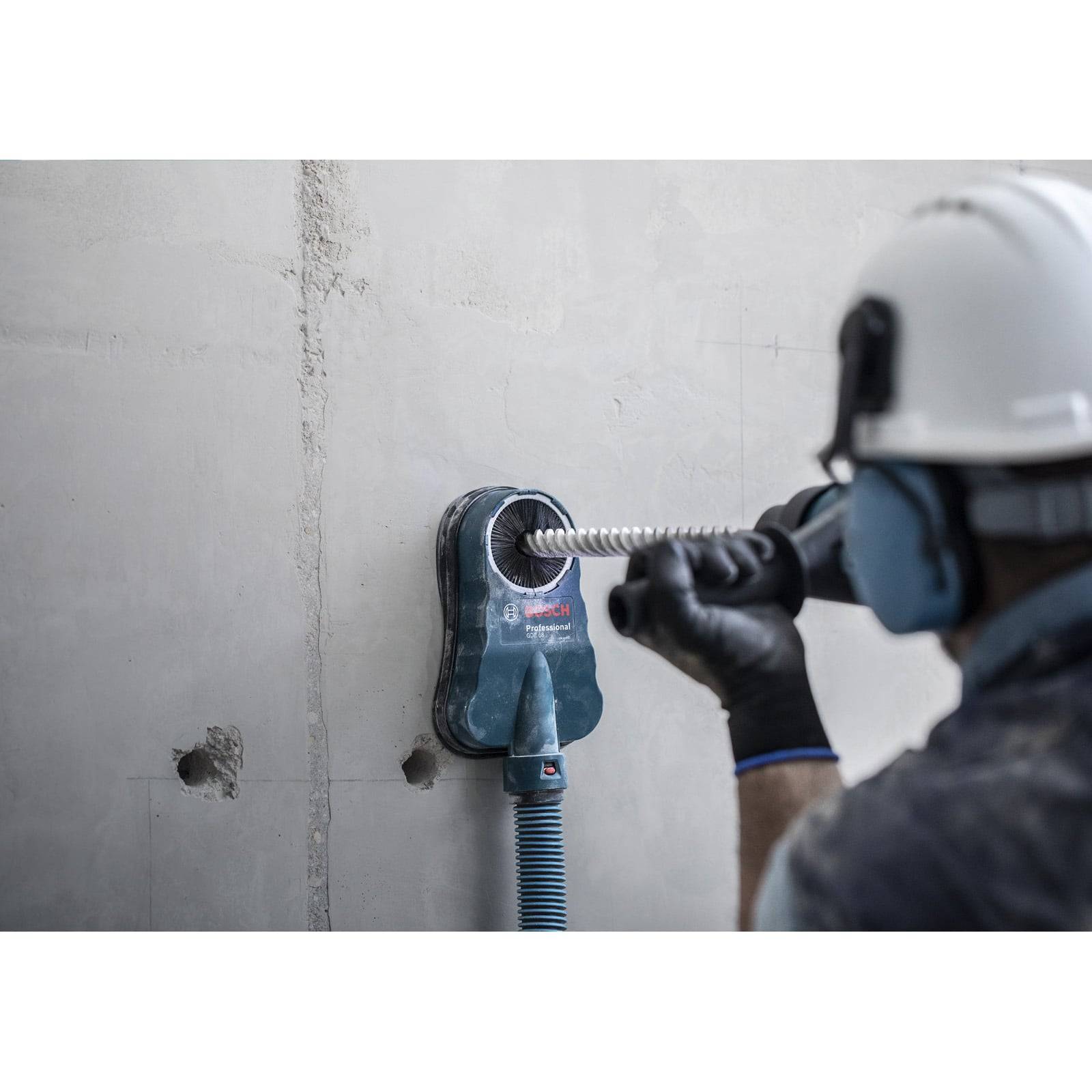 Ein Bauarbeiter mit Sicherheitsausrüstung bohrt mit einem Elektrowerkzeug mit Staubsammler-Aufsatz in eine Betonwand.