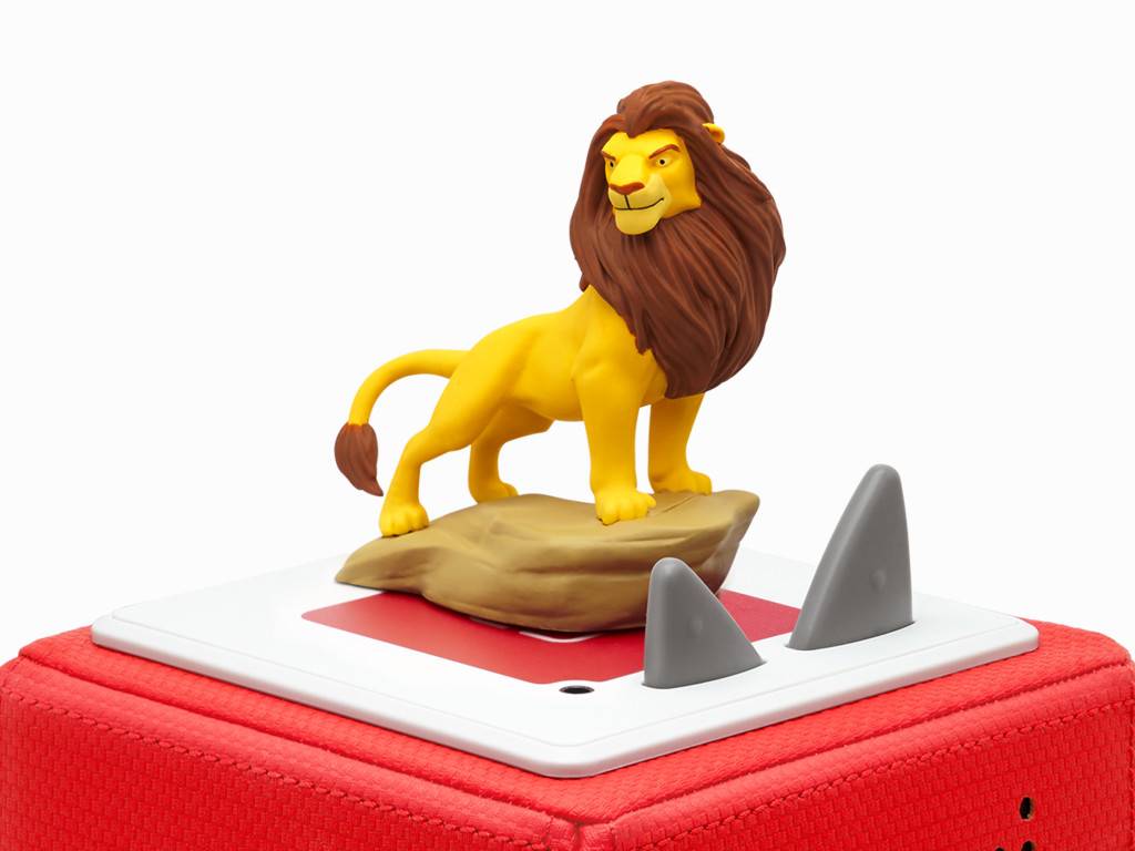 Spielzeuglöwe-Figur, die auf einem Stein steht, der sich auf einem roten Reißverschluss-Koffer mit weißem Panel und grauen Spitzen befindet, vor einem einfachen Hintergrund.