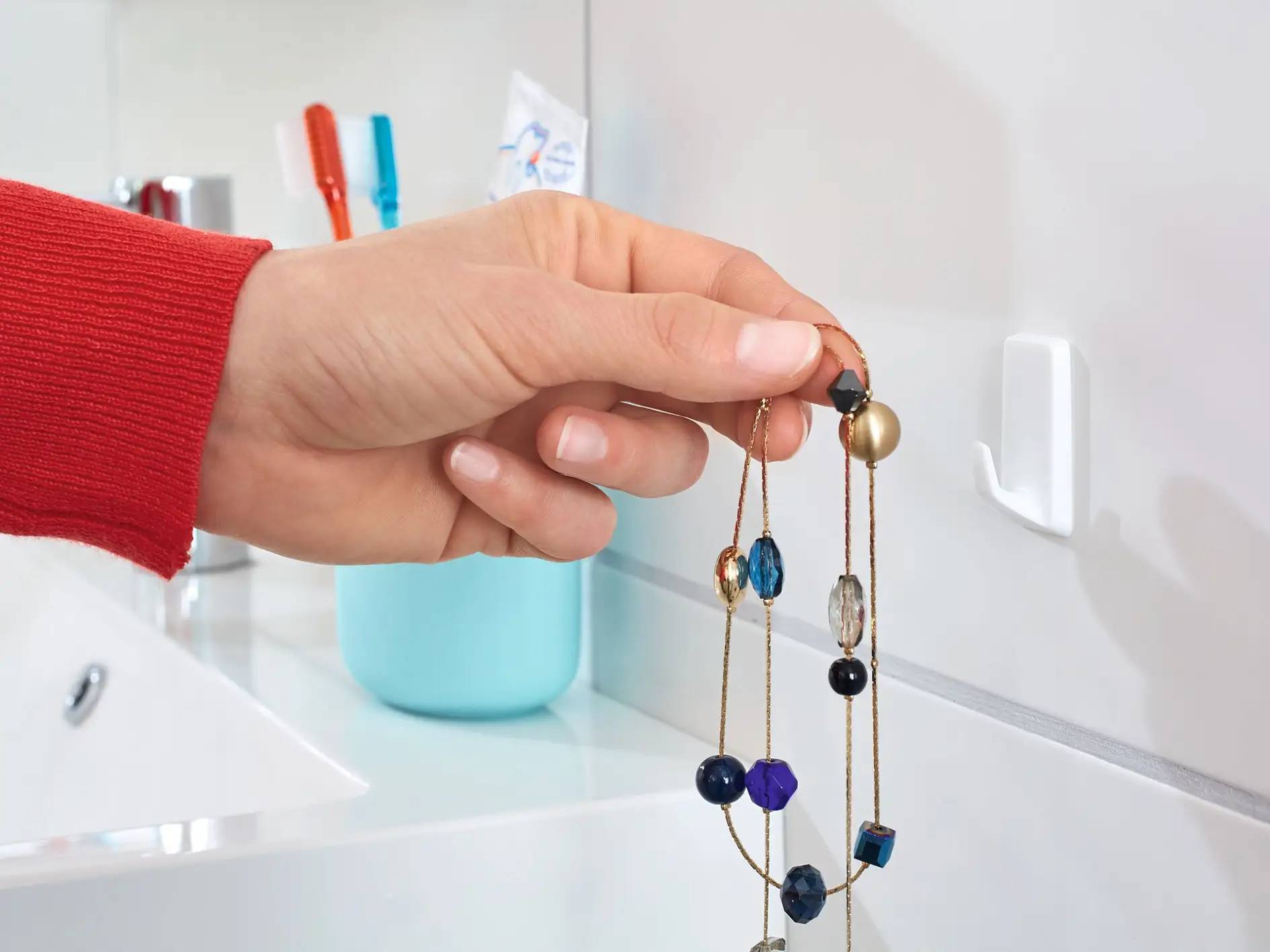 Hand, die zwei baumelnde Perlenohrringe neben einem Waschbecken und Zahnbürstenhalter im Badezimmer hält.