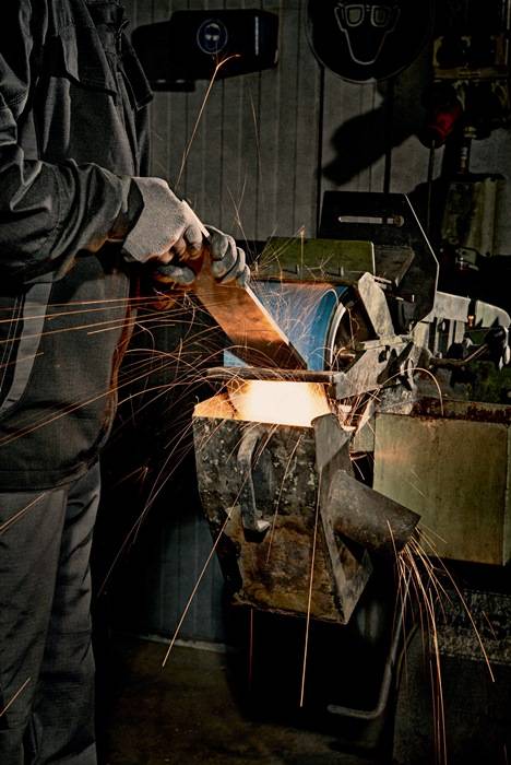 Eine Person trägt Schutzhandschuhe und bedient eine Schleifmaschine, wobei Funken in alle Richtungen sprühen. Die Szene spielt in einem industriellen Umfeld.