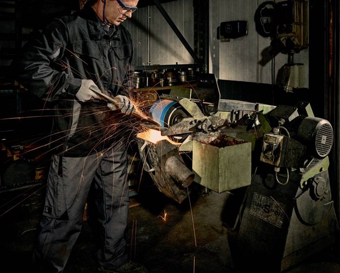 Eine Person in Schutzausrüstung bedient eine Schleifmaschine und erzeugt Funken in einer düster beleuchteten Werkstatt. Verschiedene Werkzeuge und Ausrüstungsgegenstände sind zu sehen.