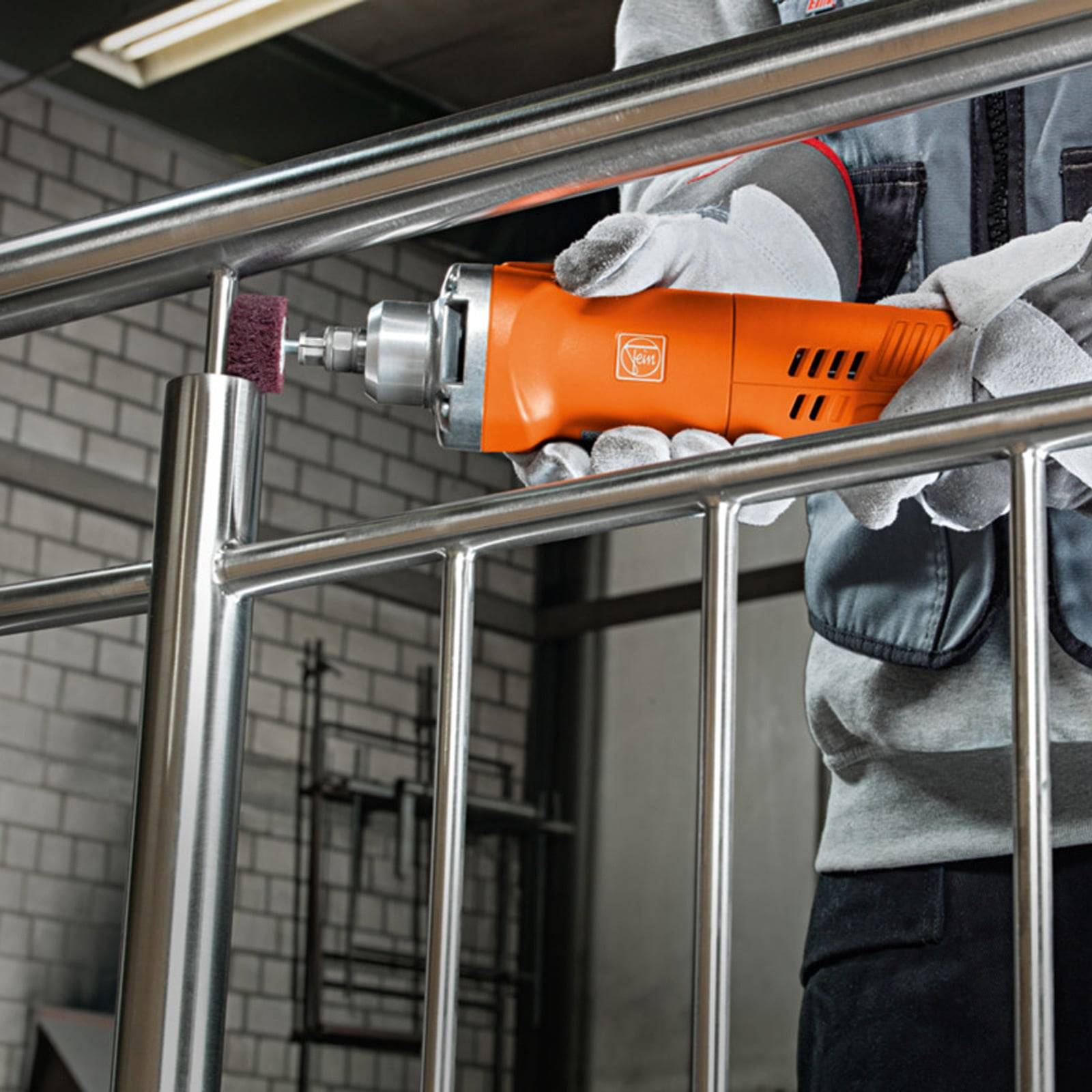 Person, der in einer industriellen Umgebung eine orangefarbene Elektrowerkzeug zur Politur eines Metallgeländers verwendet. Das Werkzeug verfügt über einen rotierenden Aufsatz zum Glätten von Kanten.