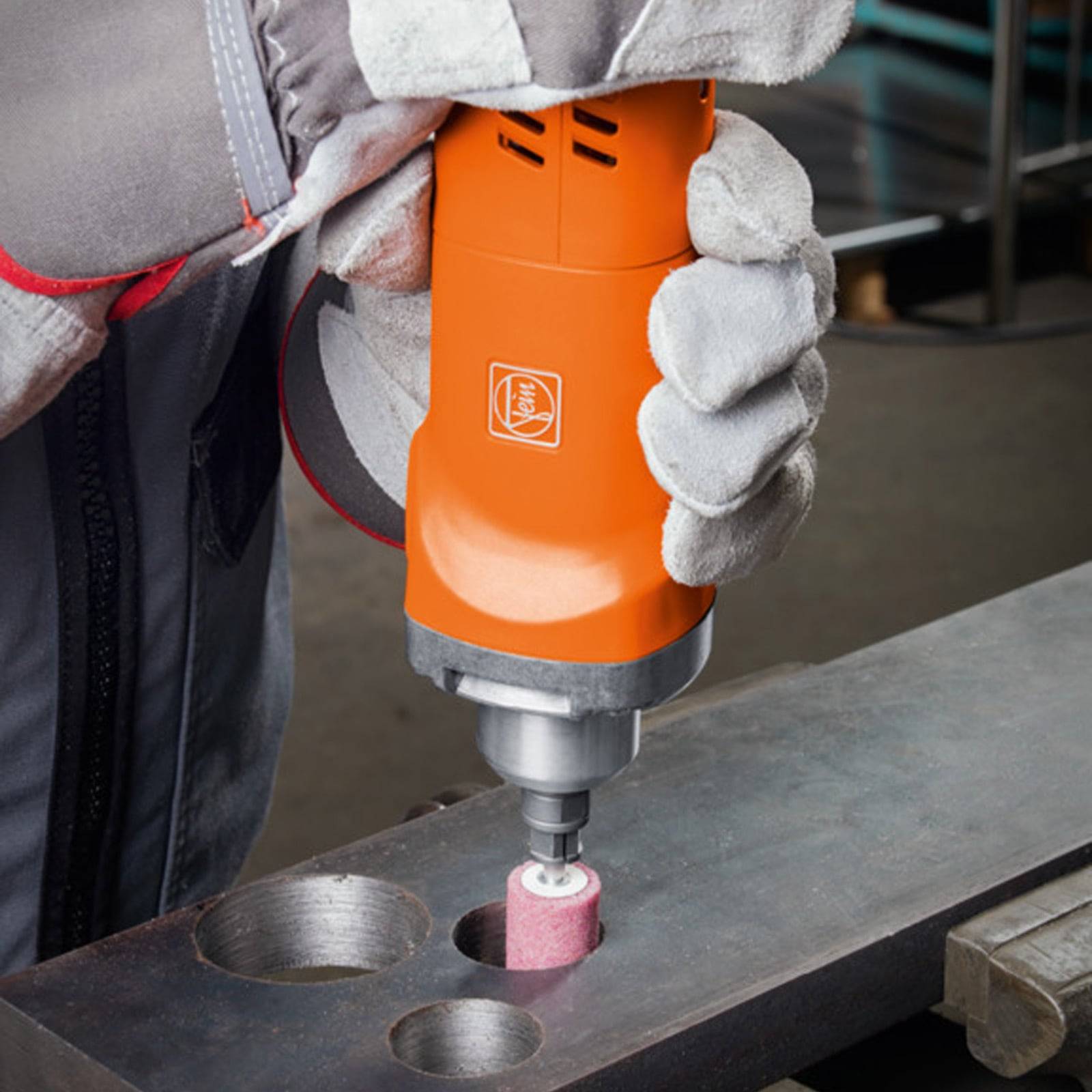 Eine Person trägt Handschuhe und verwendet ein orangefarbenes Rotationswerkzeug mit einem Schleifaufsatz, um die Kante eines Metallstücks mit Löchern zu polieren oder zu schleifen.