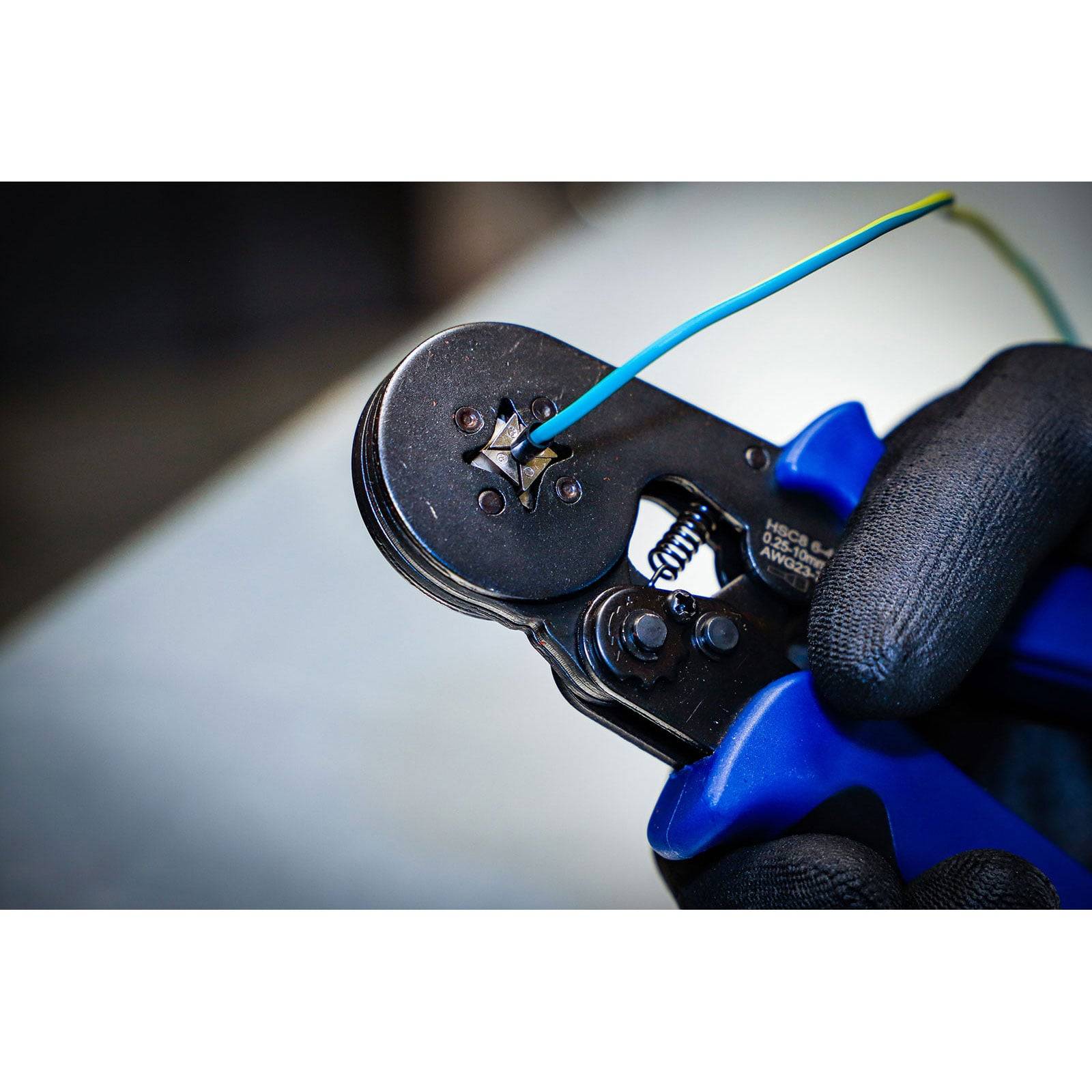 „Eine behandschuhte Hand benutzt ein Crimpwerkzeug mit blauem Griff, um eine Drahtklemme zu verbinden.