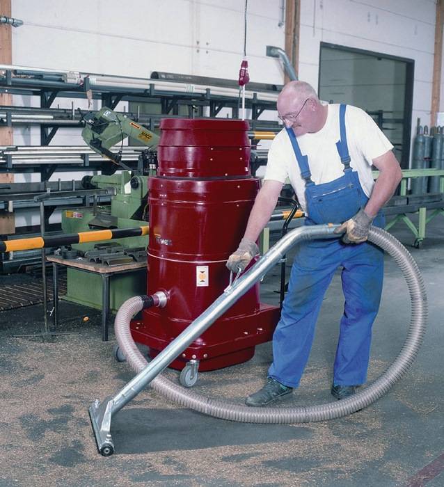 Mann, der einen industriellen Staubsauger verwendet, um Holzspäne auf einem Werkstattboden zu reinigen; Regale und Maschinen sind im Hintergrund sichtbar.