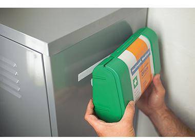 Eine Person stellt einen grün beschrifteten Sharps-Behälter in einen an der Wand montierten Metallschrank oder medizinischen Entsorgungsschlitz.