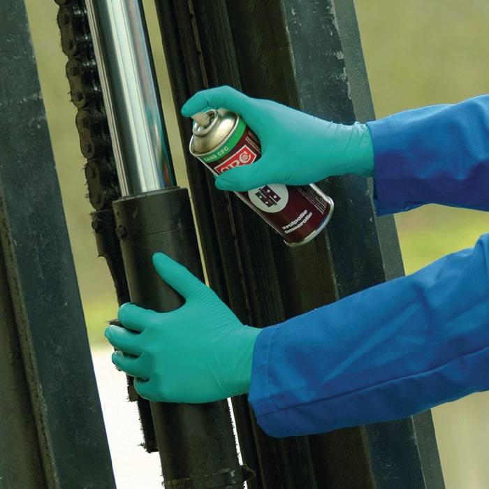Eine Person mit türkisfarbenen Handschuhen trägt Schmiermittel aus einer roten Sprühdose auf eine Metallstange und eine Kette auf, höchstwahrscheinlich für Wartungszwecke.