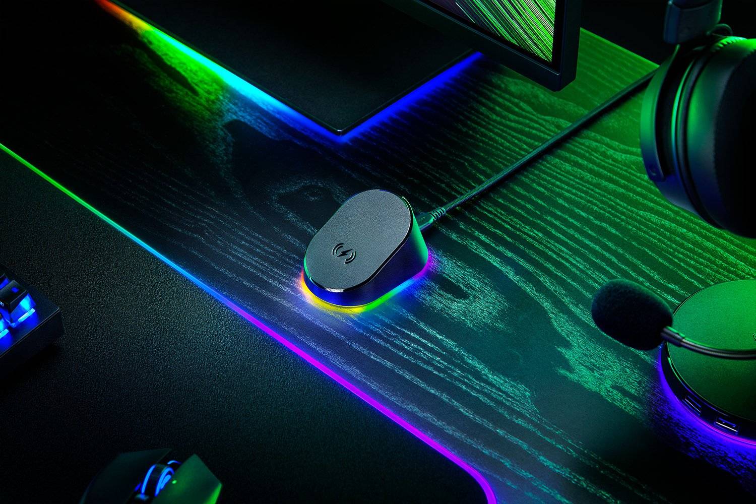 Gaming-Schreibtisch-Setup mit einem kleinen kabellosen Ladepad auf einer dunklen RGB-beleuchteten Mausmatte, plus Kopfhörer, Monitor und einer sichtbaren Tastaturkante.
