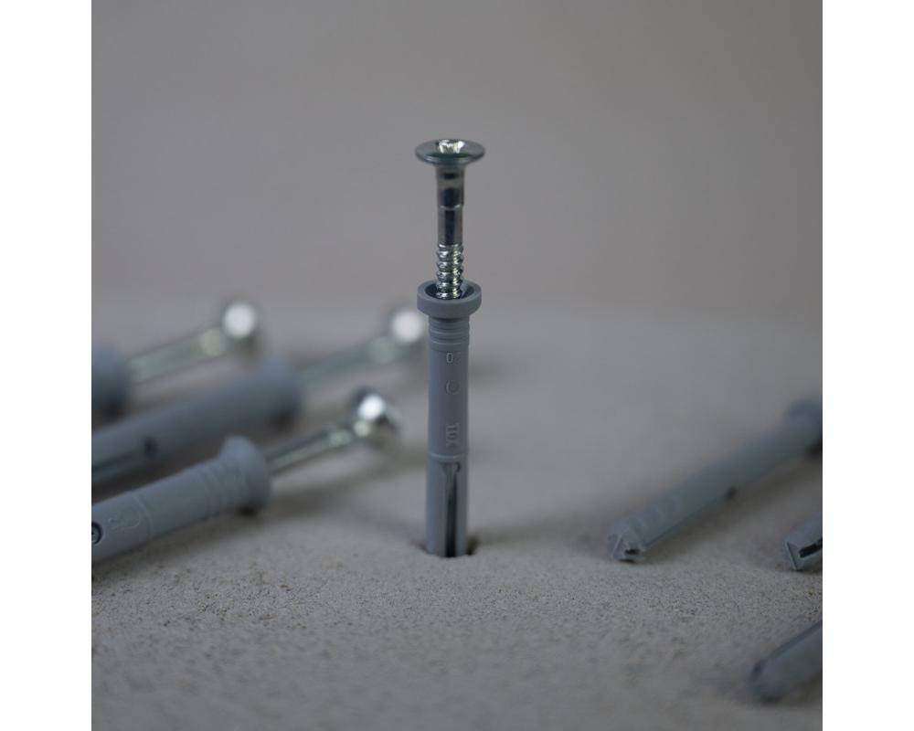 Eine Schraube wird in einen grauen Wanddübel eingefügt, der aufrecht auf einer Oberfläche steht, umgeben von anderen ähnlichen Dübeln und Schrauben, die flach liegen.