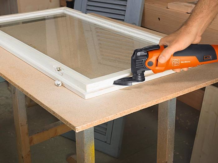 Eine Person verwendet ein orangefarbenes Multitool, um einen weißen Fensterrahmen zu trimmen, der auf einem Holztisch liegt.