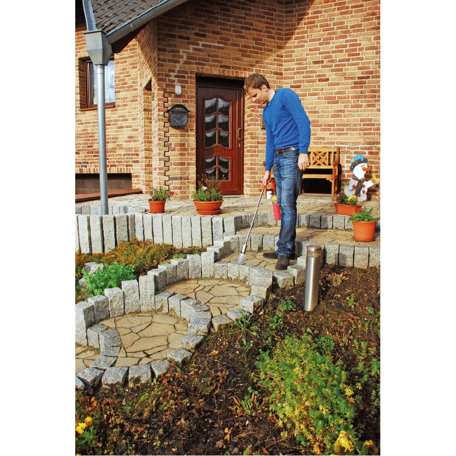 Eine Person steht auf einem steinernen Gehweg in der Nähe eines Ziegelhauses und benutzt ein Werkzeug zur Pflege des Gartens, der mit sorgfältig angeordneten Pflanzen und Töpfen gestaltet ist.