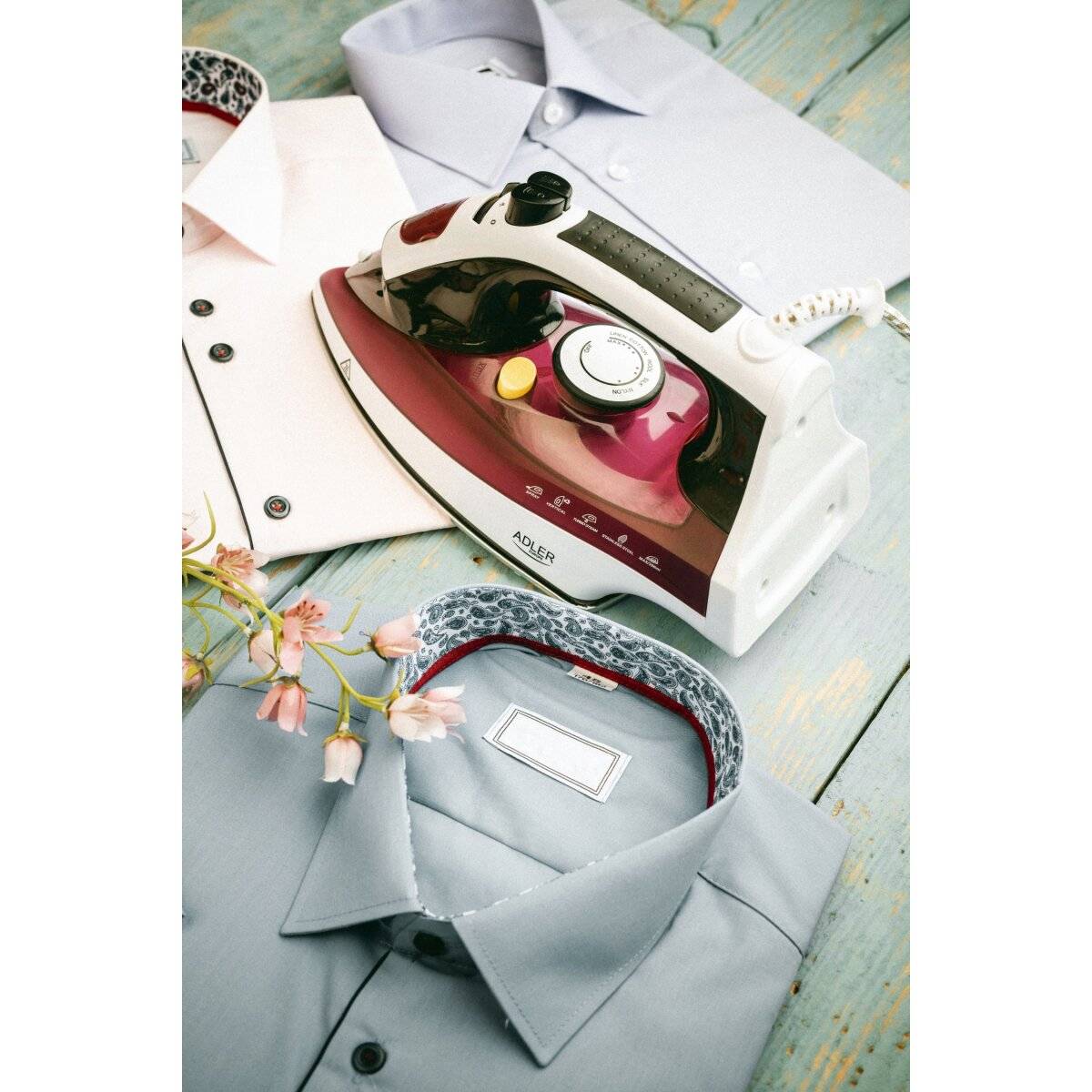 Ein Dampfbügeleisen wird auf einer Holzoberfläche neben ordentlich zusammengelegten Hemden platziert, wobei ein Hemd mit rosa Blüten in der Nähe dekoriert ist.