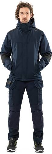 Eine Person in einem dunkelblauen taktischen Outfit mit in den Taschen verschränkten Händen vor einem schwarzen Hintergrund.