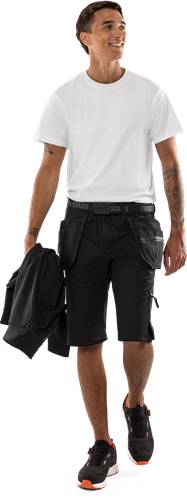 Eine Person trägt schwarze Shorts und ein weißes T-Shirt und hält eine gefaltete schwarze Jacke in ihrer linken Hand.