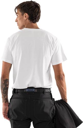 Eine Person, die ein weißes T-Shirt und schwarze Hosen trägt, mit einem sichtbaren Tattoo an einem Arm, die vor einem einfarbigen Hintergrund steht.