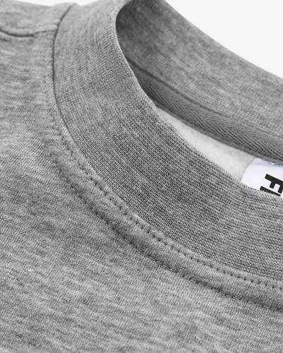 Nahaufnahme eines grauen Rundhals-Sweatshirts mit Fokus auf die Textur des Gewebes und Stickdetails. Ein teilweise sichtbares Kleidungsetikett ist zu erkennen.