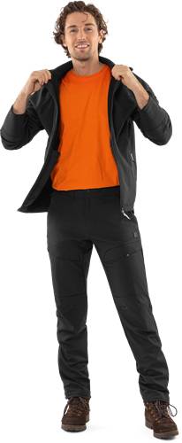 Eine Person mit einem leuchtend orangefarbenen Shirt unter einer schwarzen Jacke und schwarzer Hose, von der Taille abwärts aufgenommen.