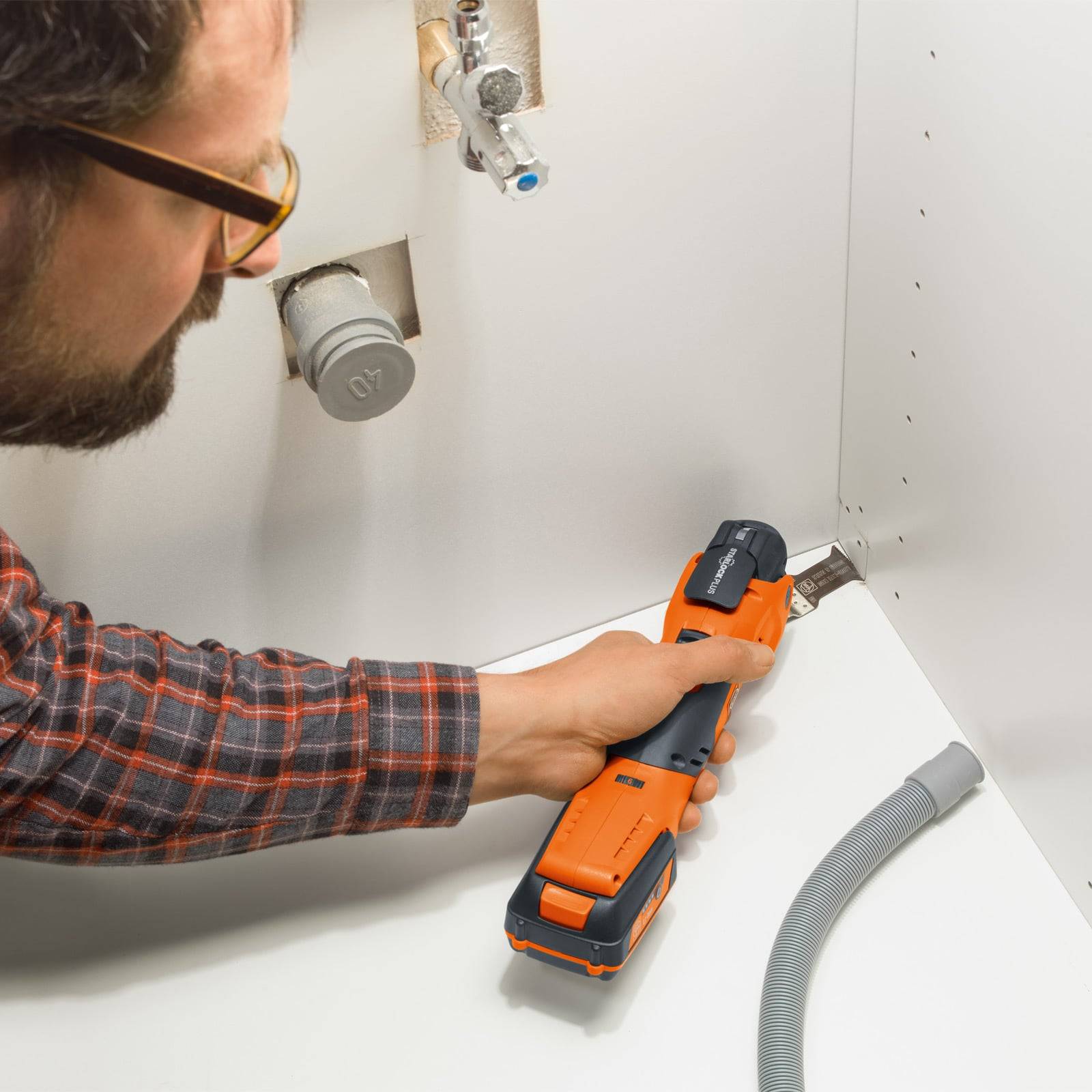 Eine Person verwendet ein Multitool, um ein Panel unterhalb eines Waschbeckens zu trimmen, wobei Klempnerarmaturen im Hintergrund sichtbar sind.