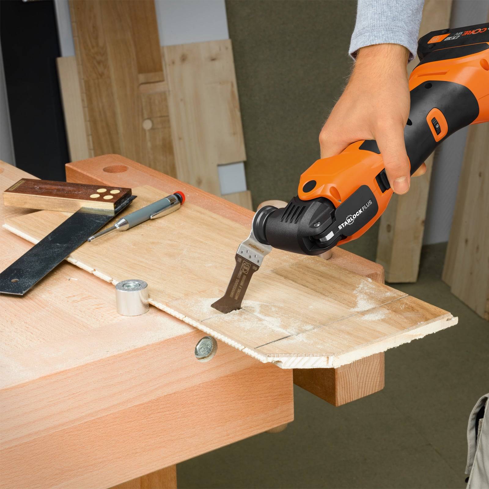 Eine Person benutzt ein orange-schwarzes oszillierendes Multifunktionswerkzeug, um ein Holzbrett auf einer Werkbank zu schleifen, umgeben von anderen Holzbearbeitungswerkzeugen.