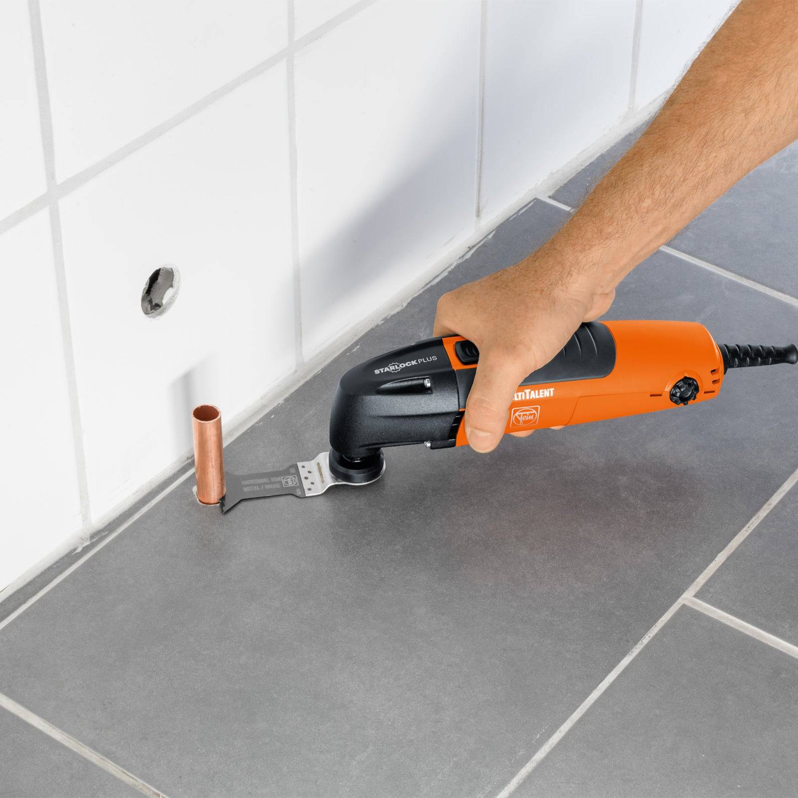 Ein Multitool mit einer oszillierenden Klinge schneidet ein Kupferrohr in der Nähe einer gefliesten Wand und eines gefliesten Bodens.