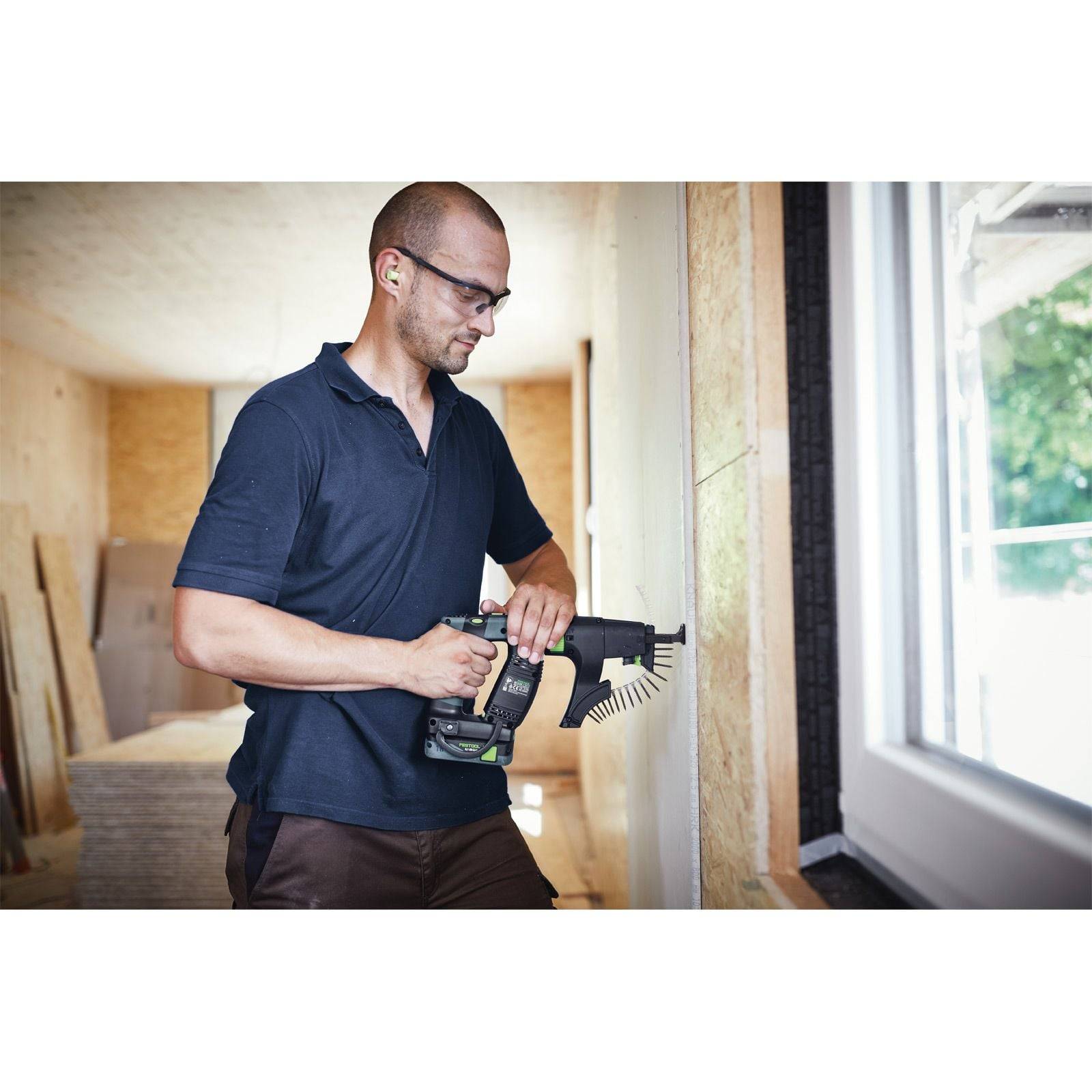 Eine Person mit Gehörschutz verwendet eine kabellose Bohrmaschine an einer Holzwand in einem hell erleuchteten Raum mit Paneelen und großen Fenstern.