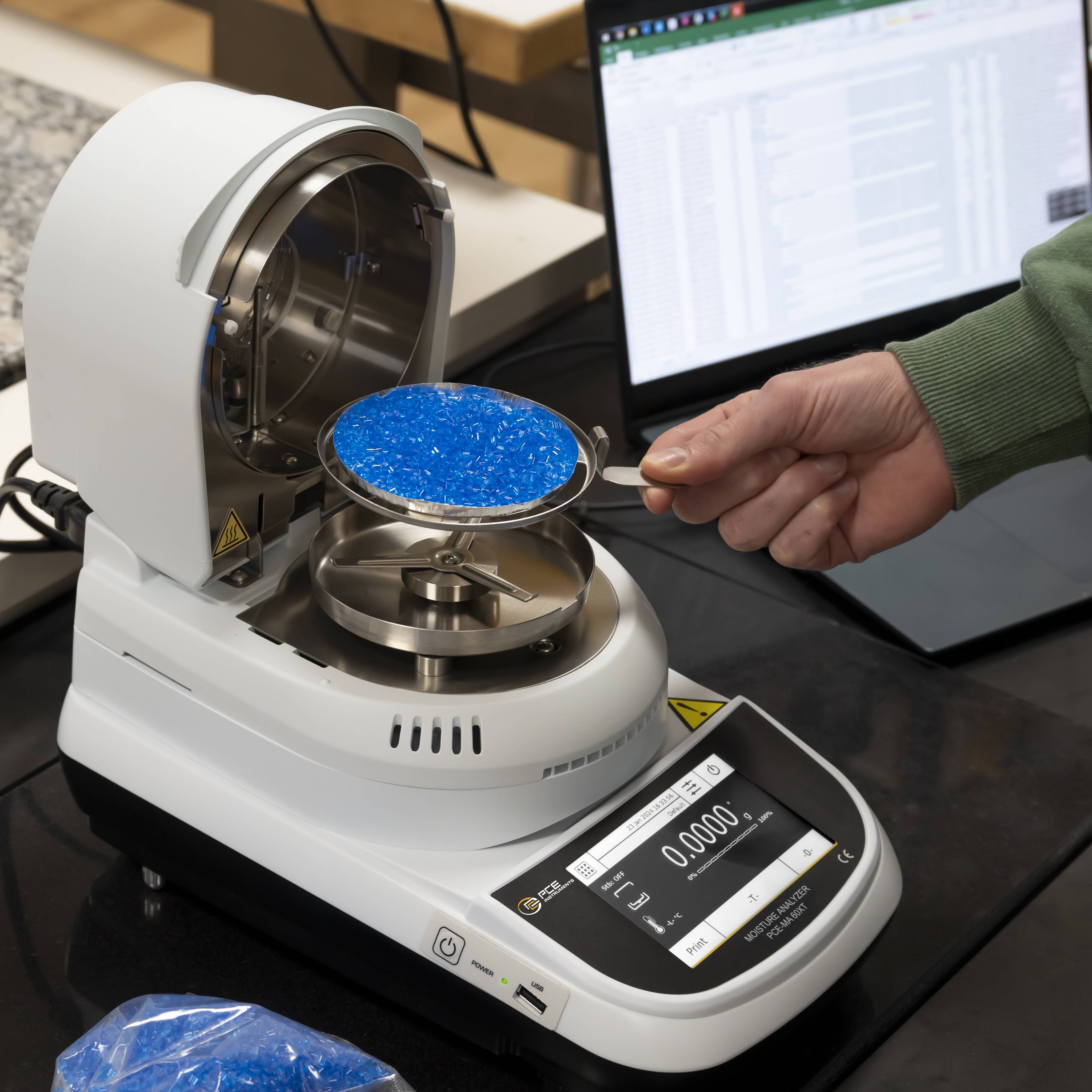 Eine Person stellt eine Schale mit blauen Granulat in ein Laborgerät zur Analyse, wobei im Hintergrund ein Laptop Daten anzeigt.