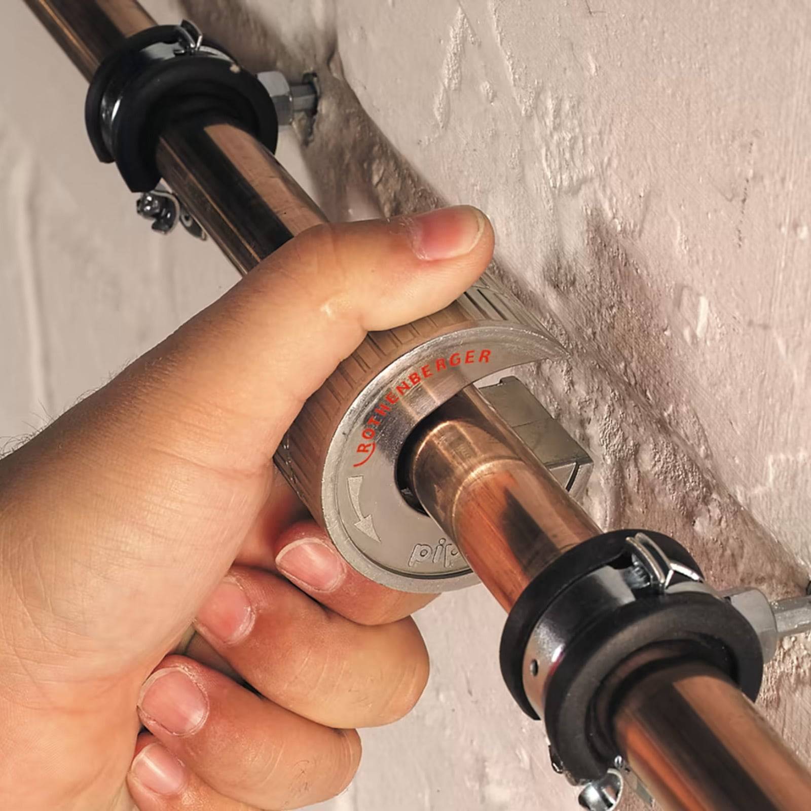 Eine Person zieht eine Kupferrohrverbindung mit einem Rohrzange fest. Die Verbindung ist an einer Wand befestigt, was auf Sanitär- oder Wartungsarbeiten hindeutet.