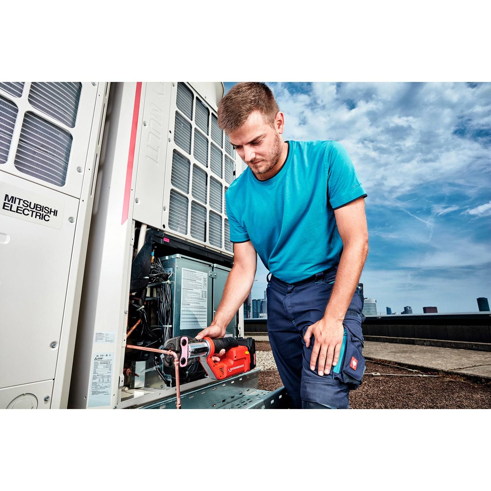 Ein Techniker in einem blauen Hemd und Arbeitskleidung repariert eine Klimaanlage auf einem Dach unter einem teilweise bewölkten Himmel.