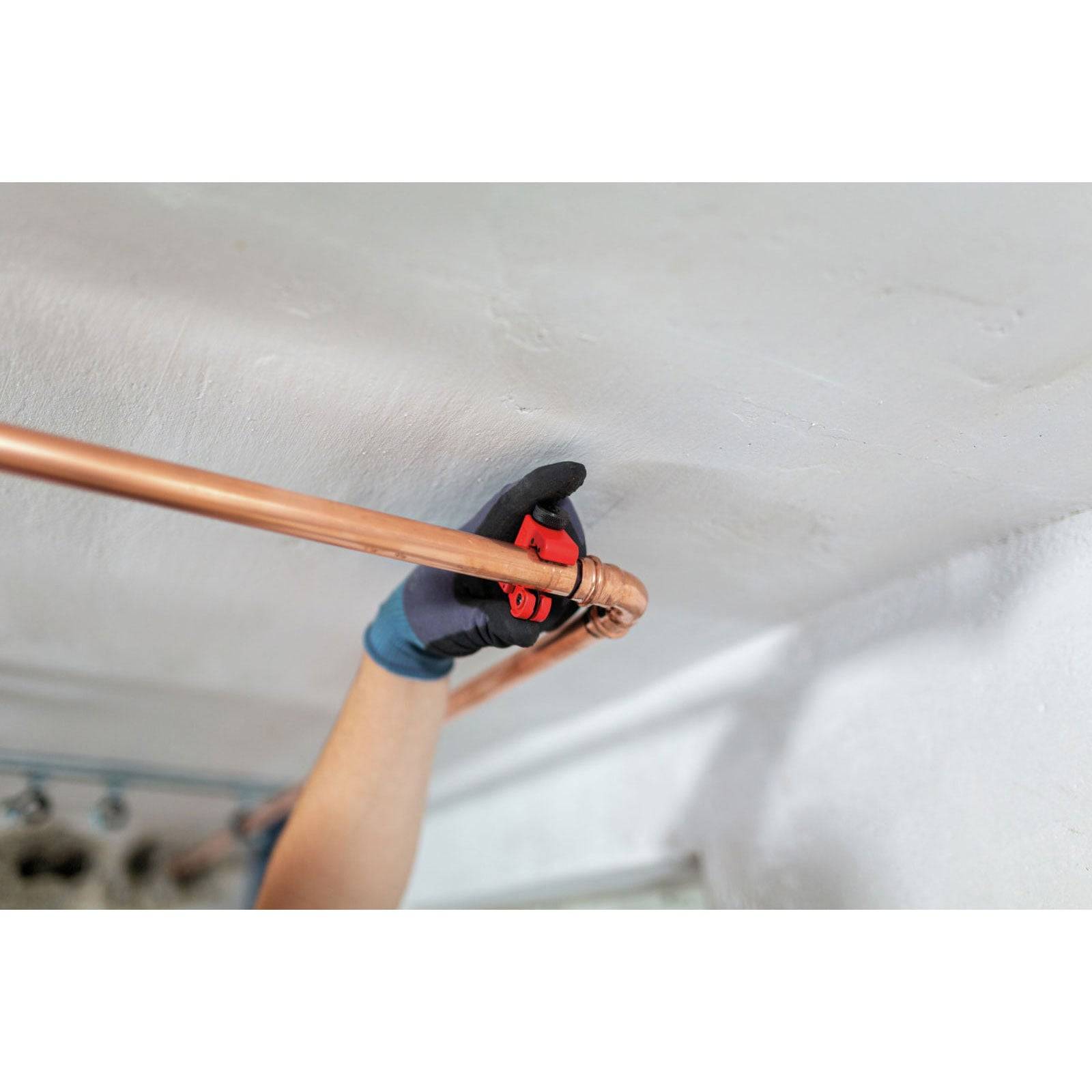 Eine Person verwendet einen Rohrabschneider, um ein Kupferrohr vor einer weißen Decke zu durchtrennen und trägt dabei Schutzhandschuhe.