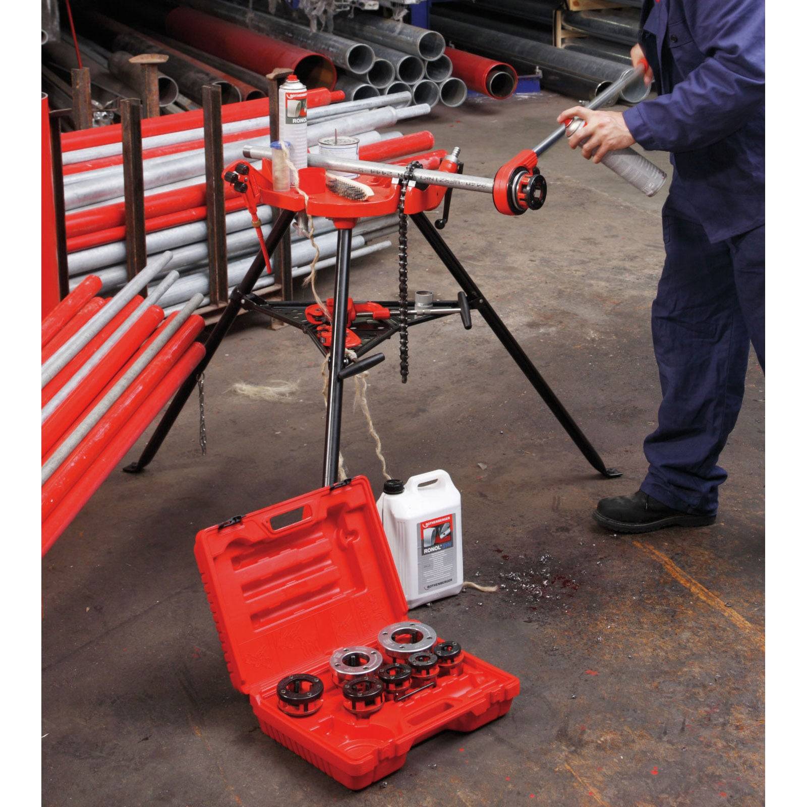 Ein Arbeiter bedient eine Rohrgewindemaschine. Rote Rohre und Werkzeuge sind verstreut umher. Eine rote Werkzeugkiste und eine Ölkanne sind auf dem Boden zu sehen.