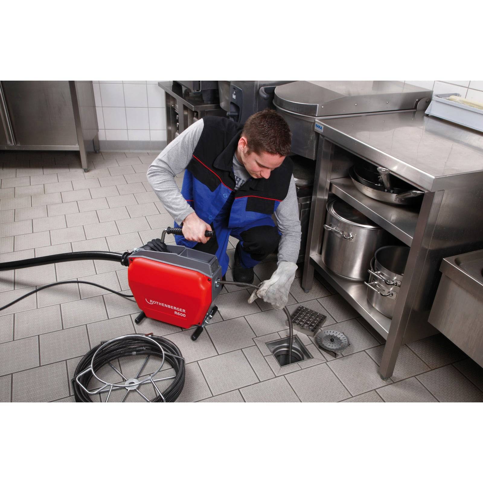 Eine Person in Uniform bedient eine Rohrreinigungsmaschine, um einen verstopften Küchenboden-Abfluss zu reinigen, umgeben von Edelstahlausrüstung.