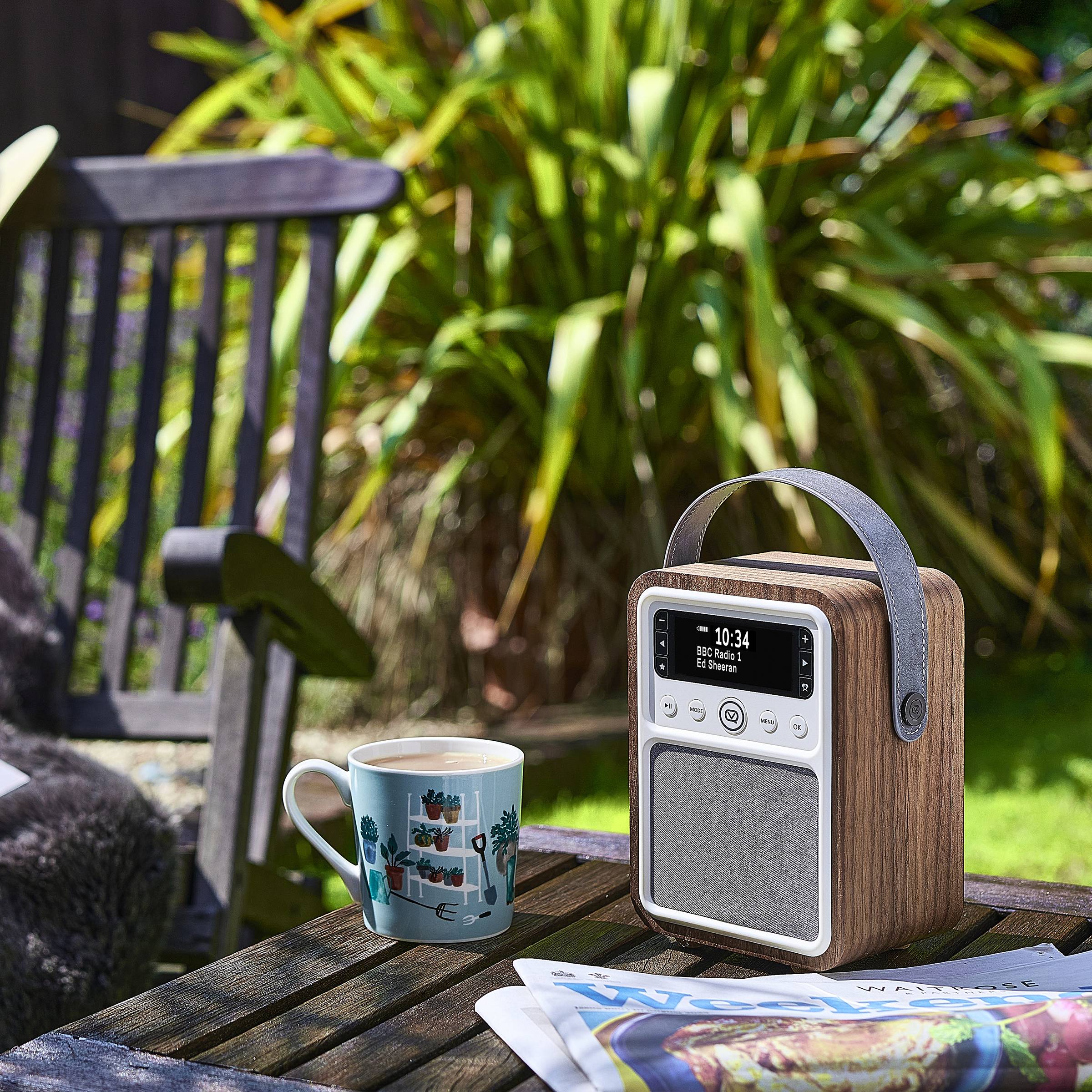 Ein Radio sitzt auf einem hölzernen Außentisch neben einer Tasse und einer Zeitung, mit einer Gartenbank und grünen Pflanzen im Hintergrund.
