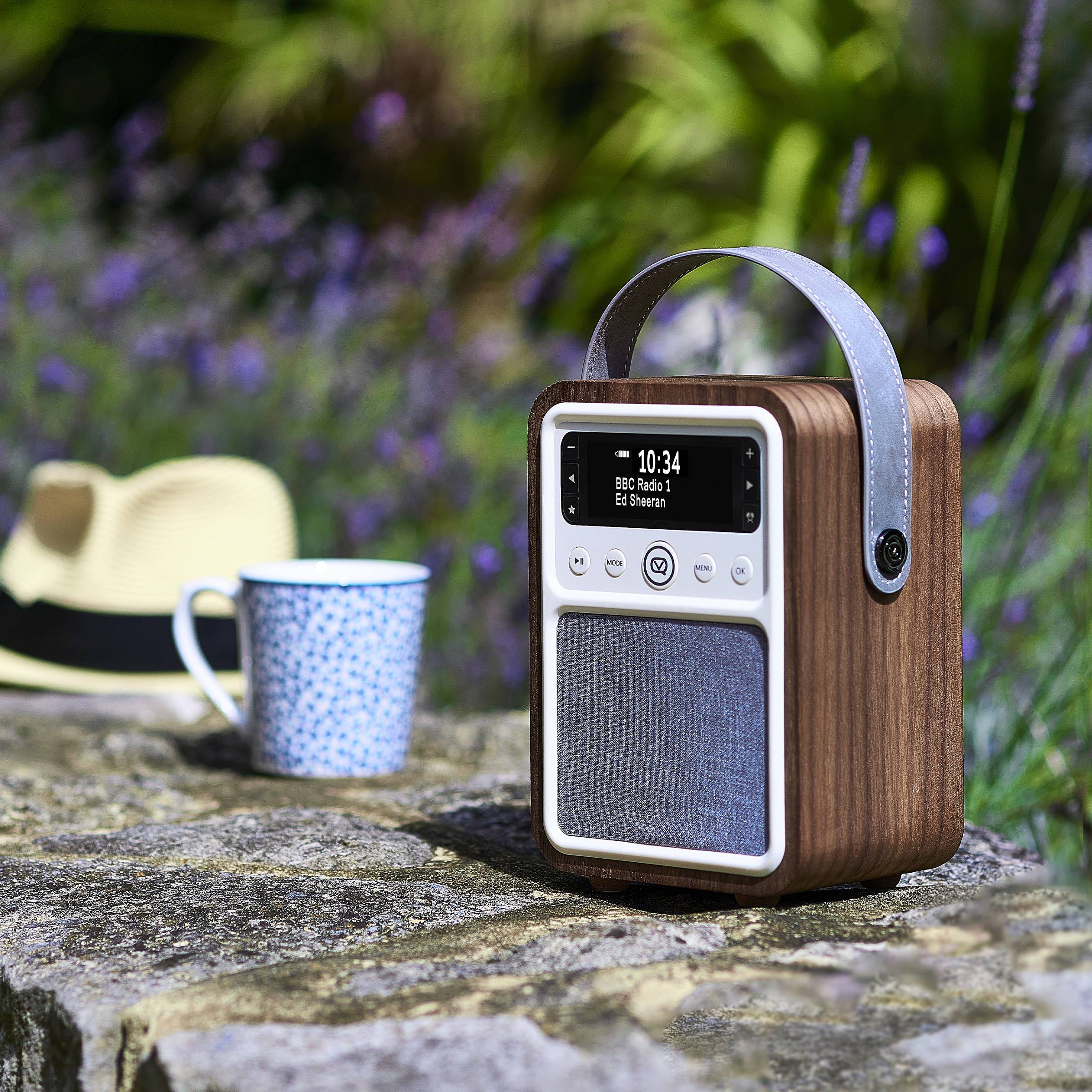 Ein tragbares Vintage-Stil-Radio mit Holzfinish und modernem Display sitzt auf einem Steinsims, umgeben von einem Hut und einer Tasse im Freien.