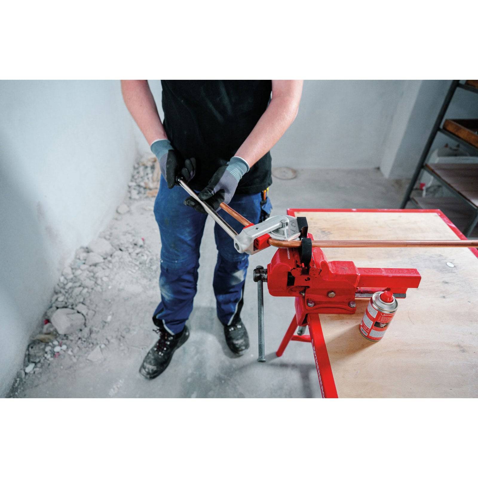Eine Person in Schutzhandschuhen bedient einen Metallrohrbieger auf einem roten Tisch mit verstreutem Schutt, was auf eine aktive Werkstattumgebung hindeutet.