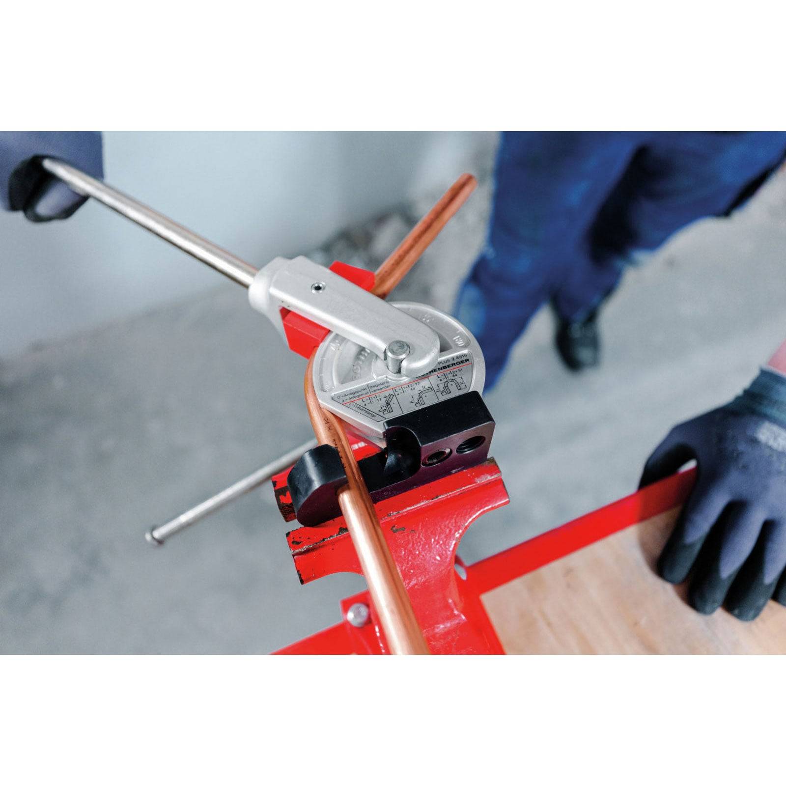 Eine Person in Handschuhen biegt ein Kupferrohr mit einem manuellen Rohrbiegewerkzeug auf einem roten Arbeitstisch und demonstriert dabei eine Klempneraufgabe.