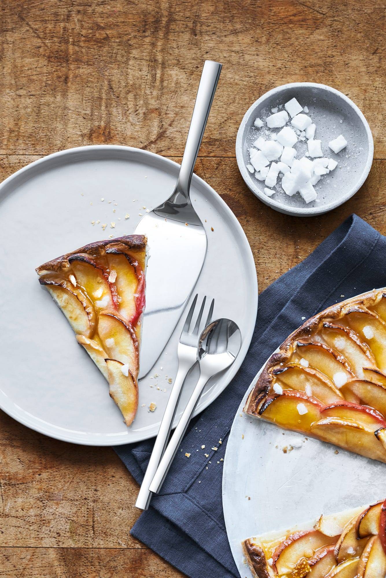 Draufsicht auf einen teilweise aufgegessenen Obstkuchen auf Tellern mit Besteck und einer Schale mit Zuckerwürfeln auf einem Holztisch.
