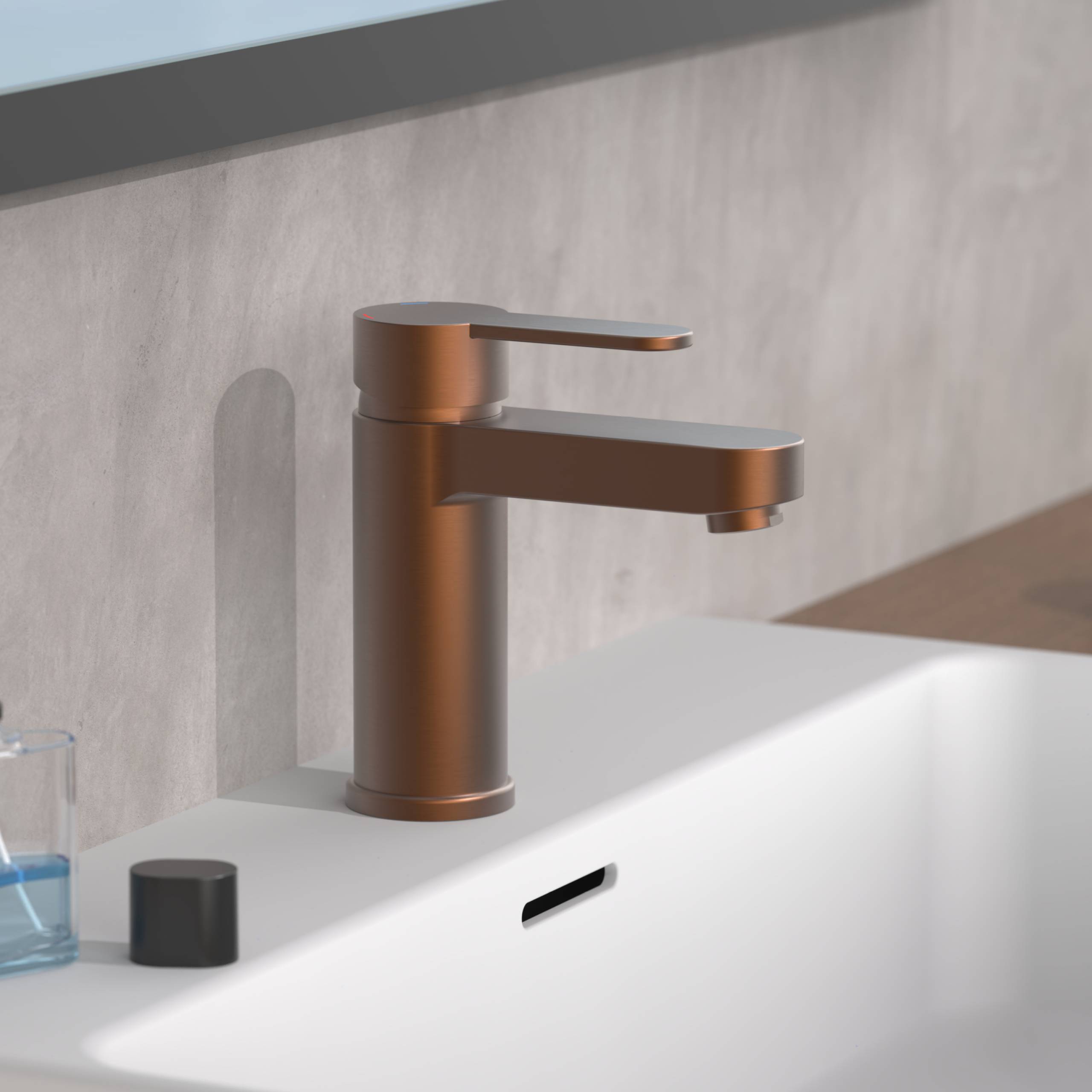 Ein moderner Bronzehahn auf einem weißen Waschbecken mit einer Rückwand mit Betontextur. Eine kleine Flasche und eine schwarze Kappe befinden sich auf der Waschbeckenkante.