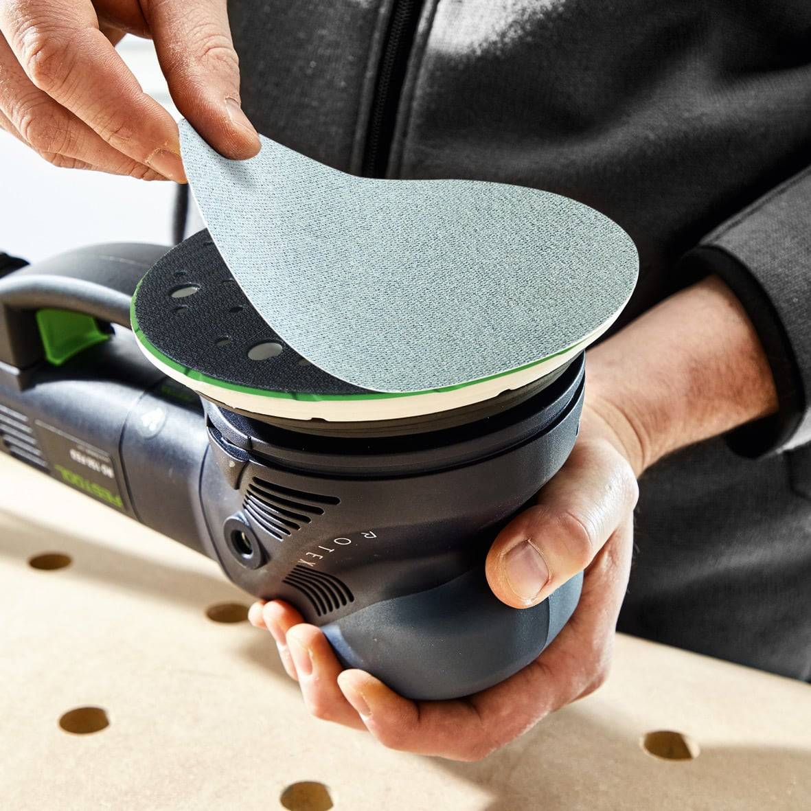 Eine Person hält einen Elektroschleiffer mit einer runden Schleifscheibe, die befestigt ist. Die Oberfläche hat mehrere Löcher, wahrscheinlich zur Staubabsaugung.