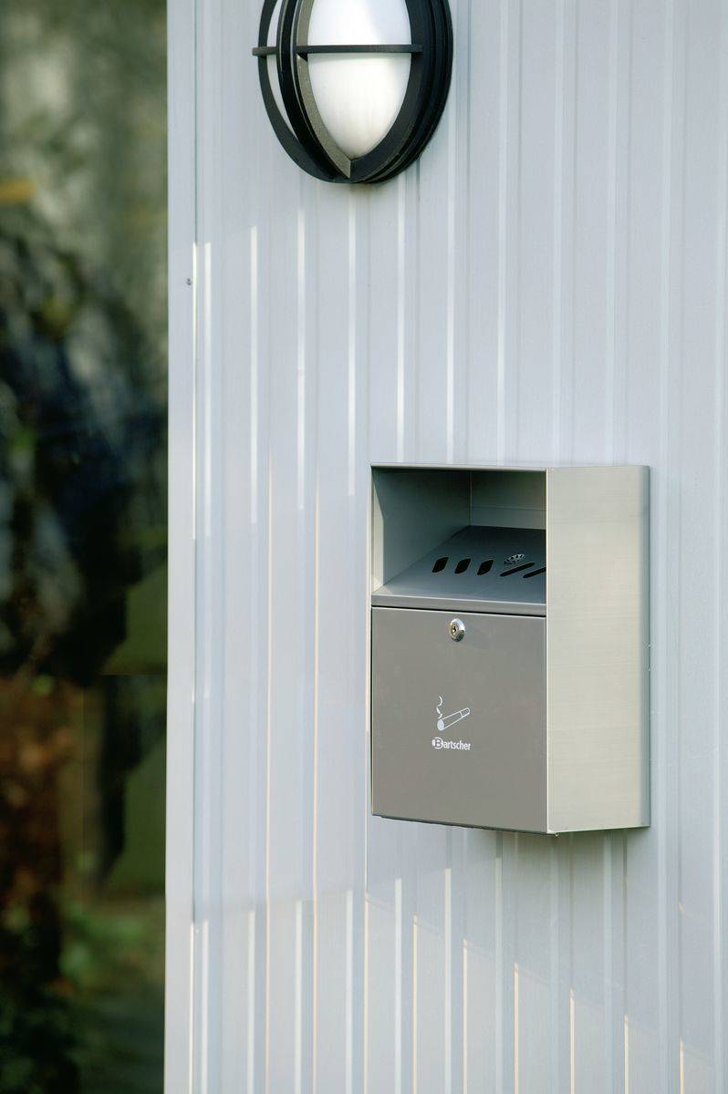 An der Wand montierter Briefkasten aus Edelstahl an einer weißen Außenwand unter einer runden Außenleuchte.