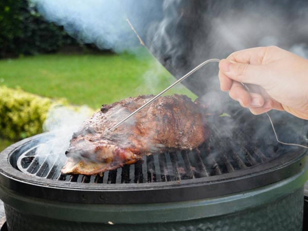 Eine Hand, die eine Fleischthermometer-Sonde in einem gegrillten Steak auf einem rauchenden Barbecue-Grill im Freien hält.
