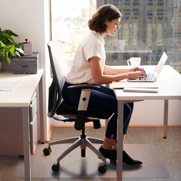 Eine Frau arbeitet an einem Laptop an einem Schreibtisch in einem hellen Büro und sitzt auf einem ergonomischen Stuhl neben einem Fenster.