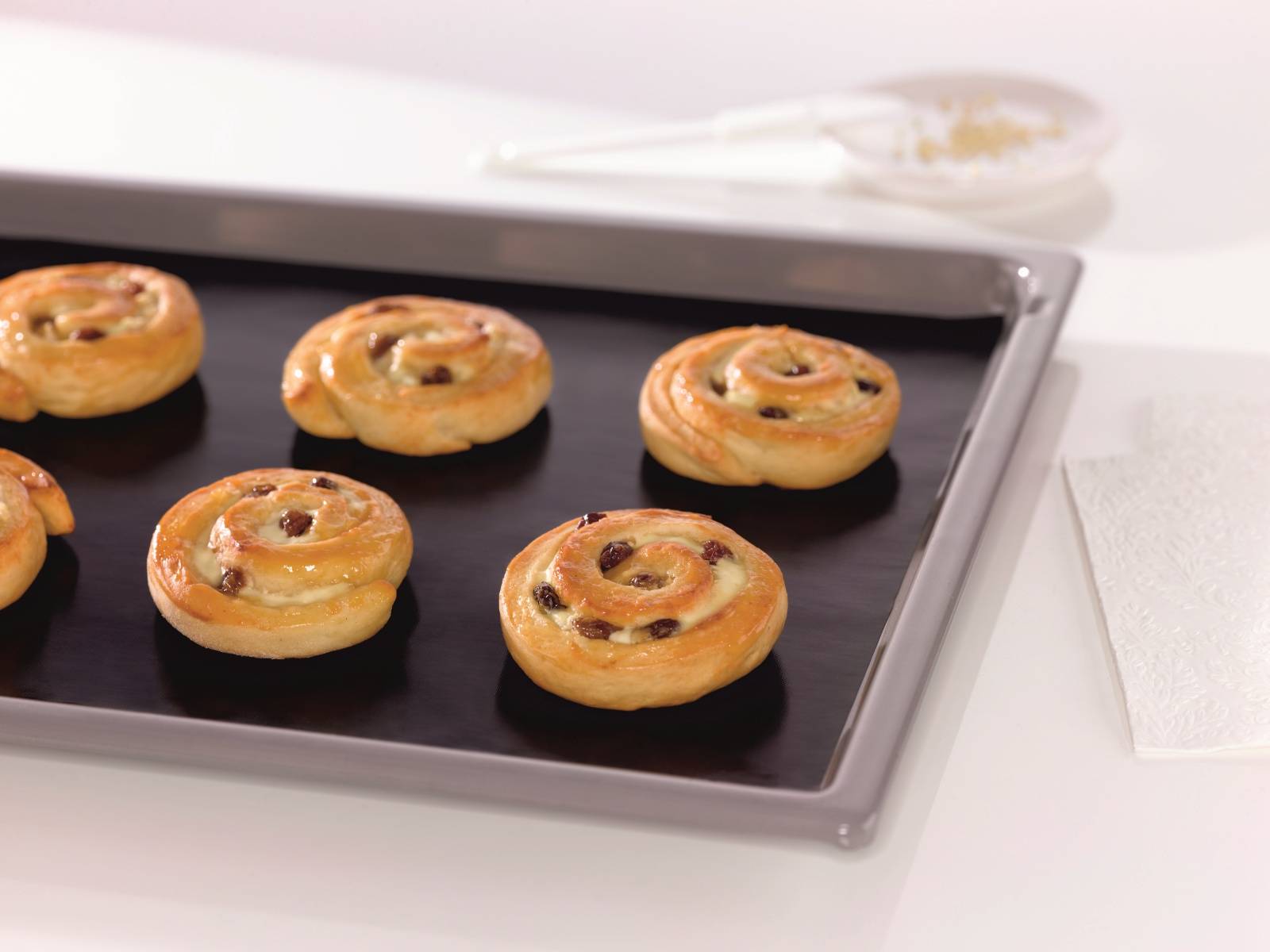 Tablett mit frisch gebackenen Spiralgebäcken mit Rosinen auf einem Backblech, das auf einer weißen Arbeitsplatte abkühlt.