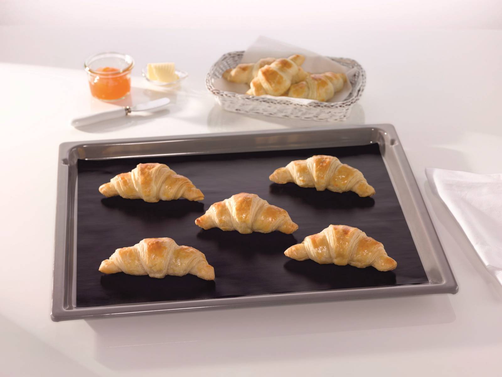 Backblech mit fünf Croissants auf einem dunklen Backblech, mit einem Korb fertiger Croissants und Marmelade und Butter im Hintergrund.